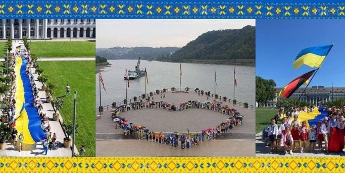 Menschen bilden ein Herzsymbol mit einer großen ukrainischen Flagge, im Hintergrund Landschaften.