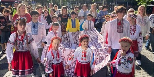 (c) Vio Miller Eine Gruppe von Kindern und Erwachsenen in traditioneller ukrainischer Kleidung feiert im Freien.